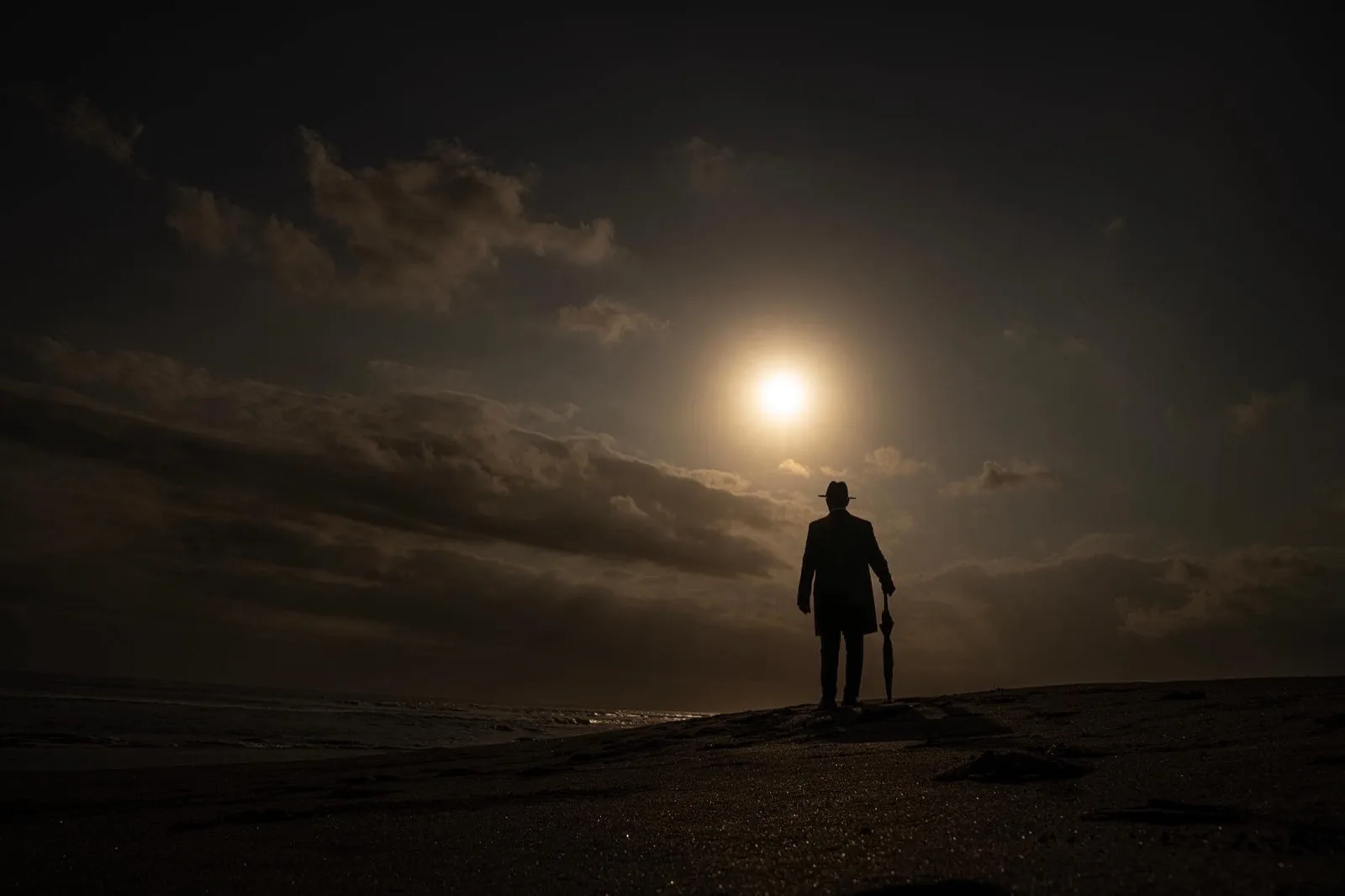 A lone traveler in a hat and coat, silhouetted on a dark shore beneath a bright moon